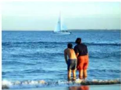 Three people standing by the sea looking at a sailboat on calm water (no text or symbols visible)