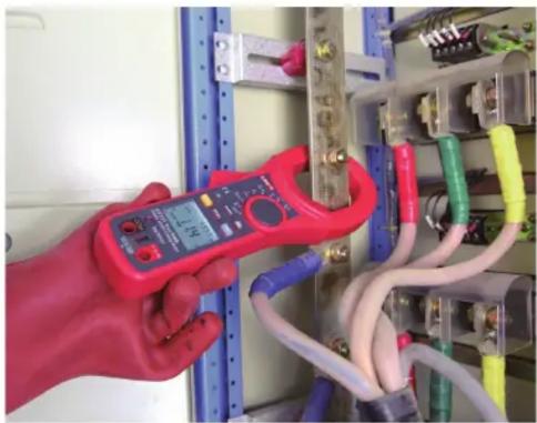 Hand holding a red handheld electrical clamp inspecting an open electrical panel with visible wiring and components (no text or symbols)