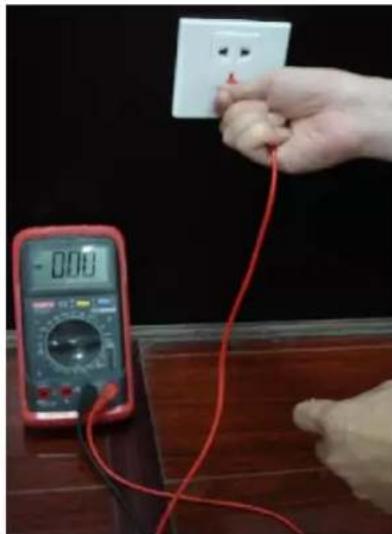 Hand holding a white electrical outlet connected to a red wire, with a digital multimeter displaying the power supply (no text or symbols visible)