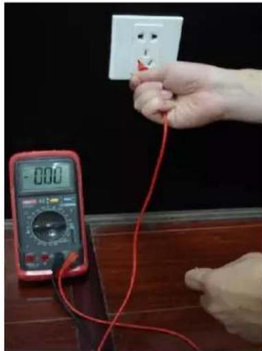 Hand holding a white electrical outlet connected to a red wire, with a digital multimeter nearby (no visible text or symbols)