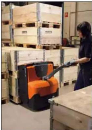 Warehouse worker operating a pallet jack in a warehouse, surrounded by stacked pallets (no visible text or symbols)