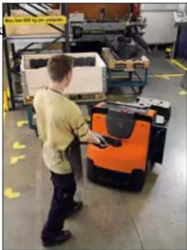 Factory worker operating a large orange and black robotic pallet in a warehouse (no visible text or symbols)