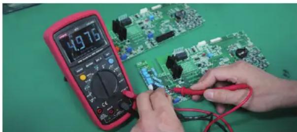 Close-up of hands using a red multimeter to test electronic circuit boards on a green workbench (no visible text or symbols)