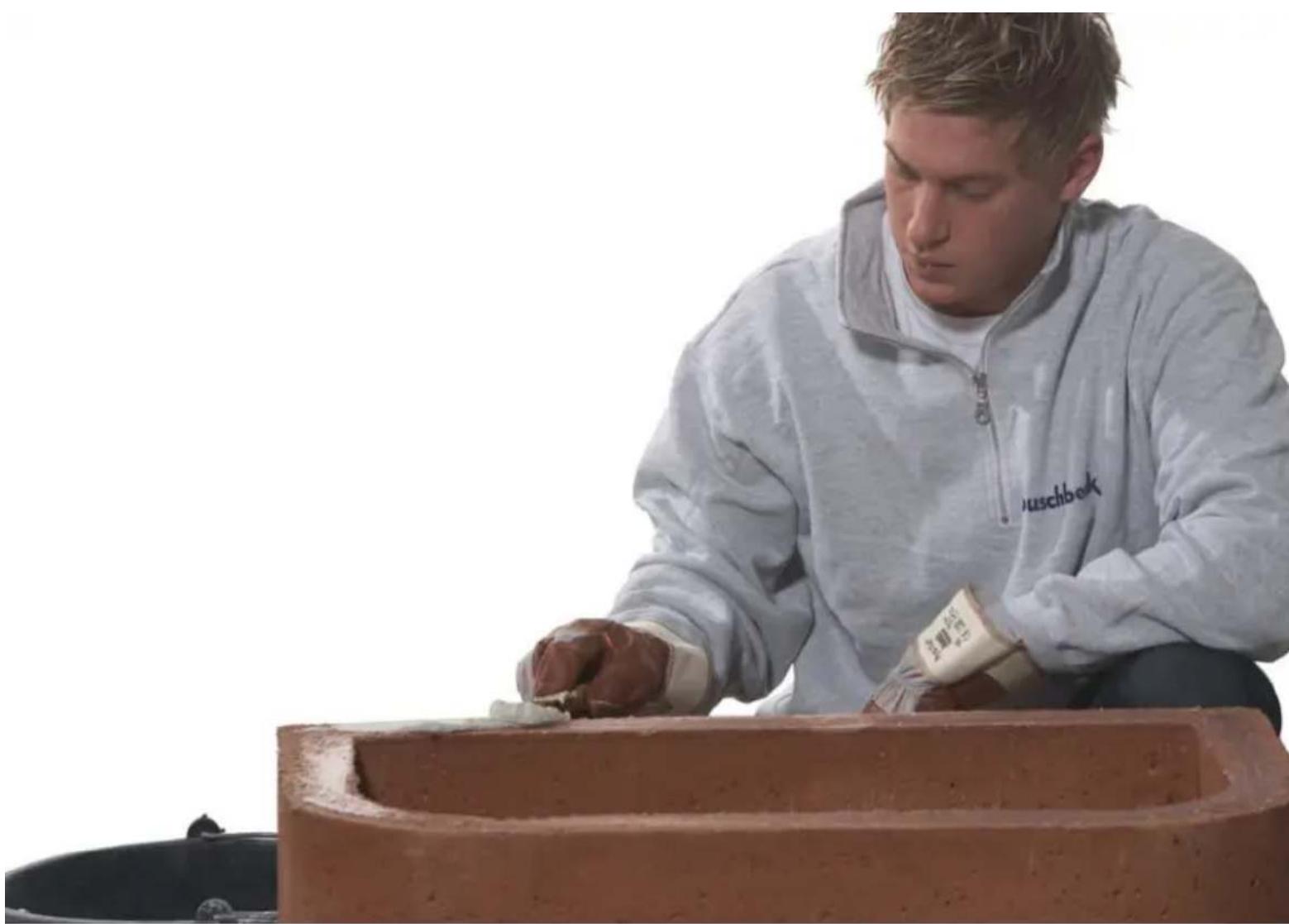 Person shaping clay on a brick structure, wearing a light jacket with 'auschbok' branding (no visible text or symbols)