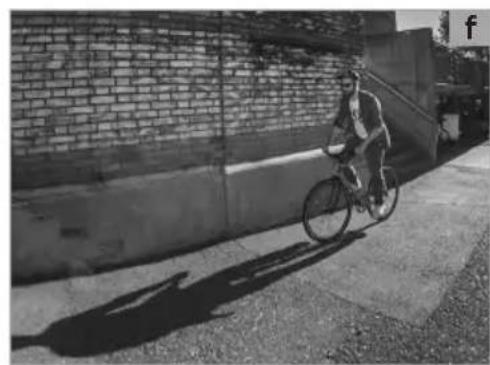 Black-and-white photo of a person riding a bicycle on a paved sidewalk beside a brick wall and concrete structure (no visible text or symbols)