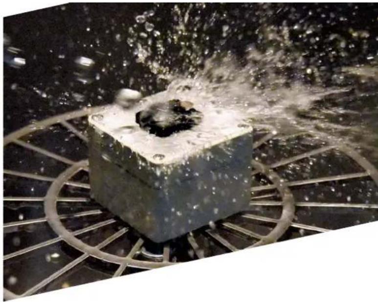 Close-up of a metallic cube with water spray and bubbles, surrounded by radial metal rings (no text or symbols visible)