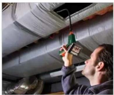 Man inspecting ceiling ductwork with a green electrical plug (no visible text or symbols)