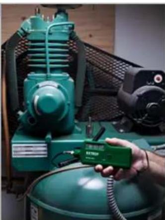 Hand holding a green electronic device labeled 'ETTRO' next to an industrial machine (no visible text or symbols on the device itself)