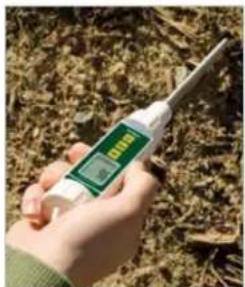 Hand holding a handheld electronic device displaying a green and black digital display with measurement markings, set against a background of dry soil.