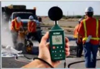 Street photo showing a hand holding a digital measuring device displaying a reading of 50.7, with workers in safety vests and hard hats in the background.