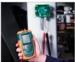 Person holding a digital multimeter in front of an electrical circuit board (no visible text or symbols)