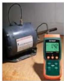 Exterior view of an electric motor on a wooden surface next to a digital handheld device displaying 142 (no visible text or symbols on the device itself)