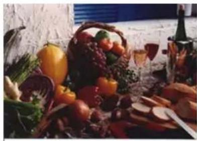 Still life arrangement of assorted fruits and vegetables on a table, including a basket of grapes, apples, and wine glasses (no text or symbols visible)