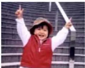 Child wearing red vest and hat, smiling with raised arms in front of a tiled wall (no text visible)