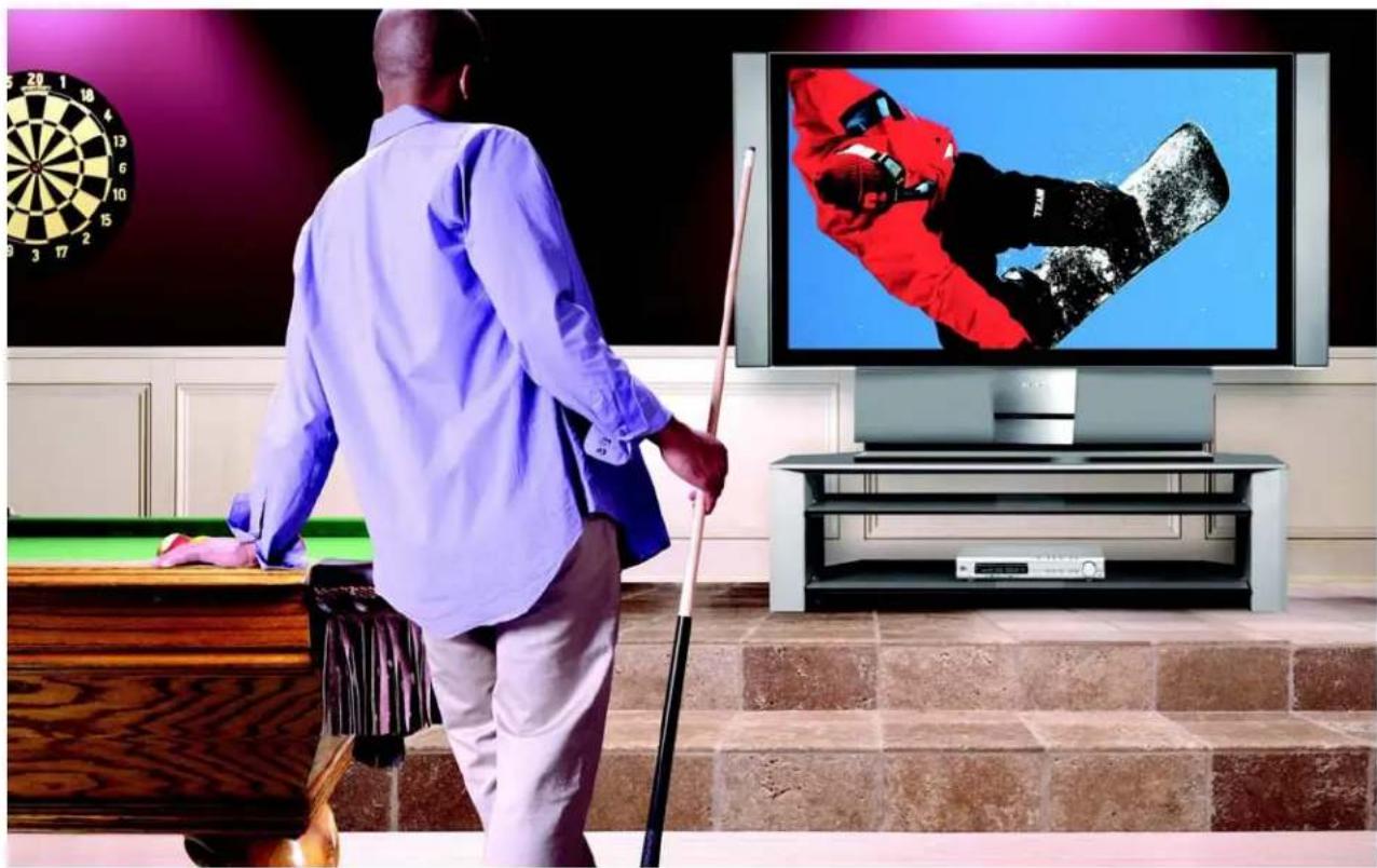 Man in blue shirt pushing cue stick toward a pool table with a dartboard, watching a TV displaying a snowy sports character (no text or symbols visible)