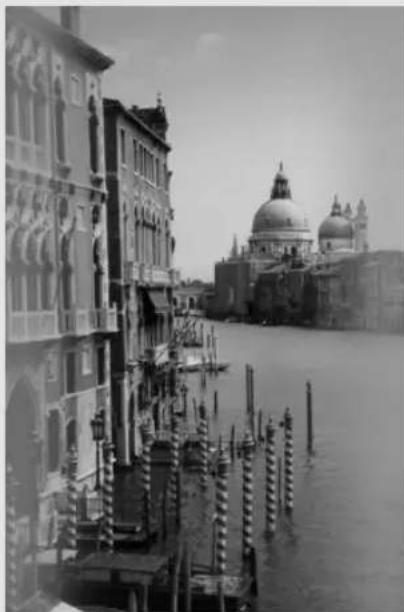Black-and-white photo of a canal scene with historic buildings and a large domed building in the background (no visible text or symbols)