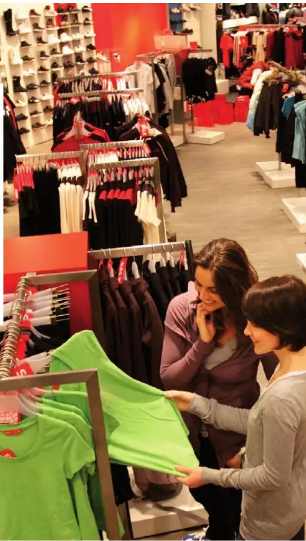 Interior of a clothing store with shoppers browsing green T-shirts on racks, surrounded by red and white racks (no visible text or signage)