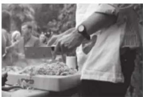 Black-and-white photo of a person preparing food outdoors, with blurred background figures and no visible text or symbols.