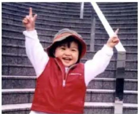 A smiling child in a red vest and hat, posing with raised fists in front of a patterned stadium (no text visible)