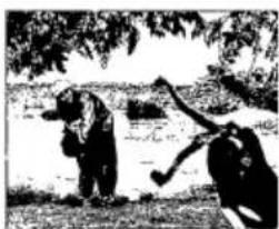 Black-and-white photo of two people near a body of water, one holding a tool, under trees (no text or symbols visible)