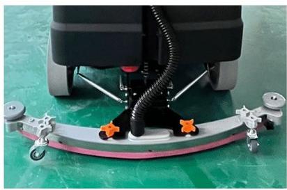 Close-up of a cleaning or cleaning robotic device with attached metal frame and orange buttons, mounted on a green tiled floor (no visible text or symbols)