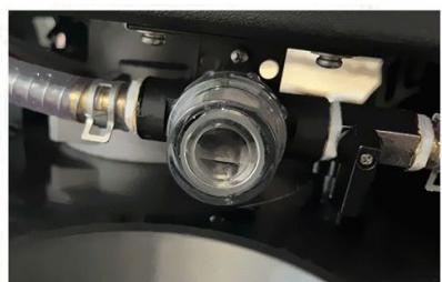 Close-up of a mechanical component with a transparent cylindrical fitting and attached tubing (no visible text or symbols)