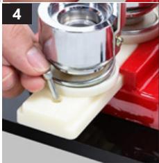 Close-up of a hand using a metal tool to press or press a small cylindrical object on a white base, with a red background (no visible text or symbols)