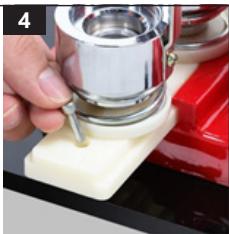 Close-up of a hand using a metal stand mixer on a white base, with red equipment in the background (no visible text or symbols)
